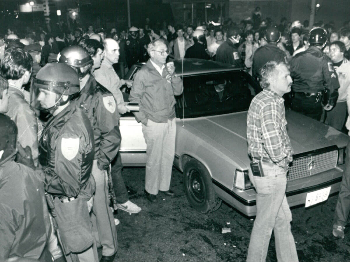 Police officers and a crowd gather around a car in a night-time street scene.