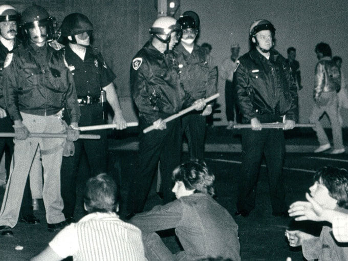 Police officers with helmets and batons confront seated individuals at night.