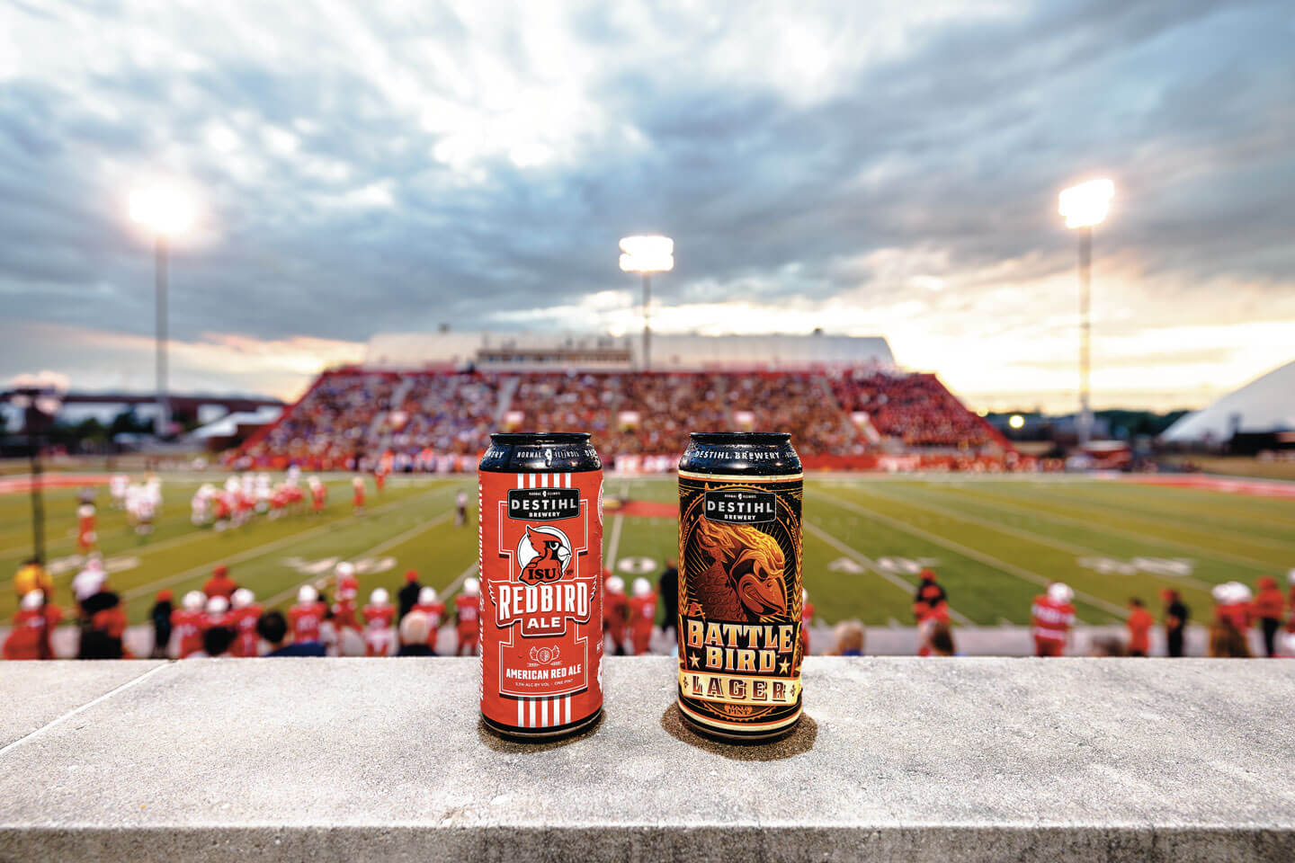 Two beer cans on a concrete ledge with a football stadium in the background.