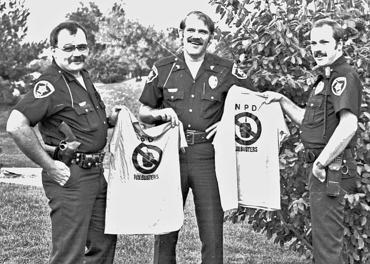 Three uniformed police officers outdoors, each holding a shirt with 'NPD - FUNBUSTERS' and a crossed-out keg symbol.