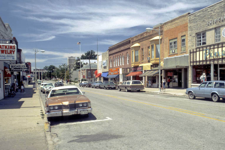 A vintage street scene with parked cars and small shops under a partly cloudy sky.