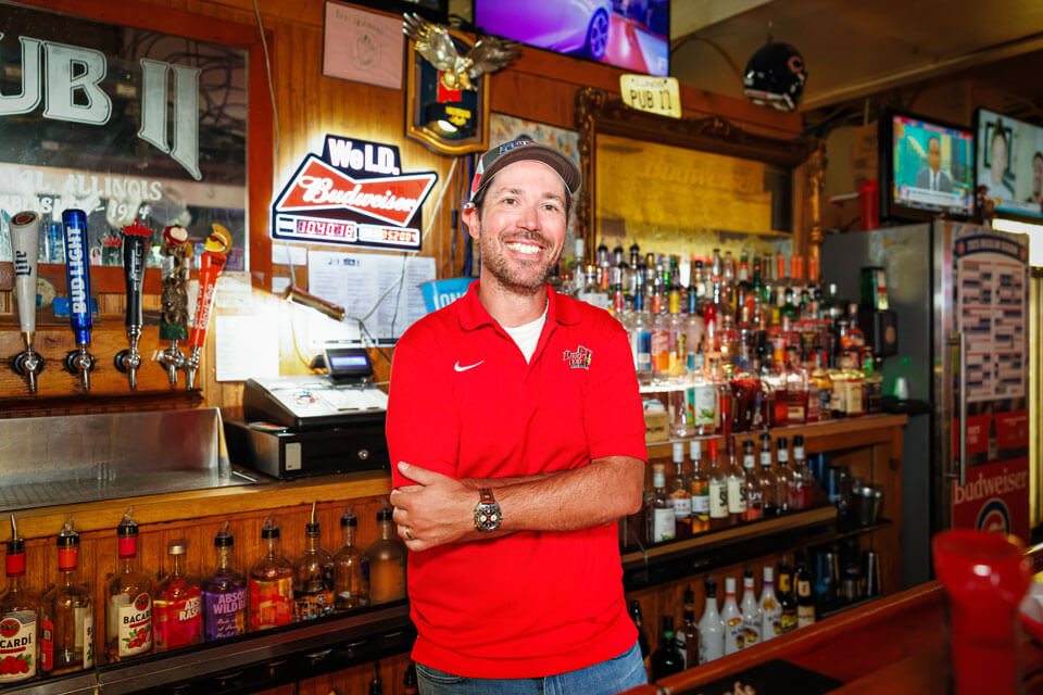 Man standing behind a bar with liquor bottles and beer taps, smiling at the camera.