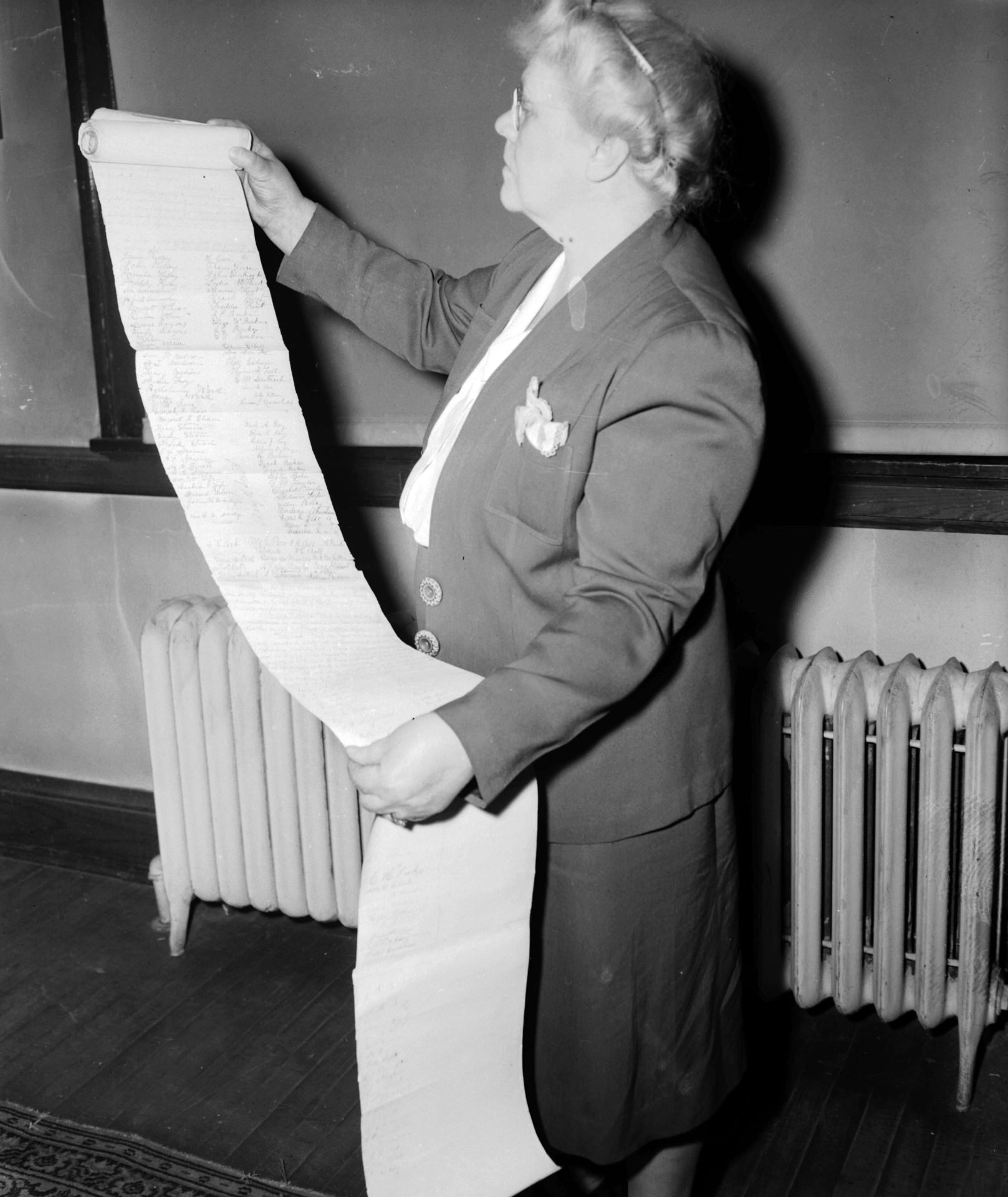 An elderly woman examines a long scroll of handwritten text indoors.