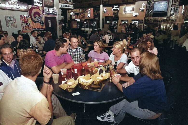 A group of people socializing around a table in a bar with neon signs and sports decor in the background.