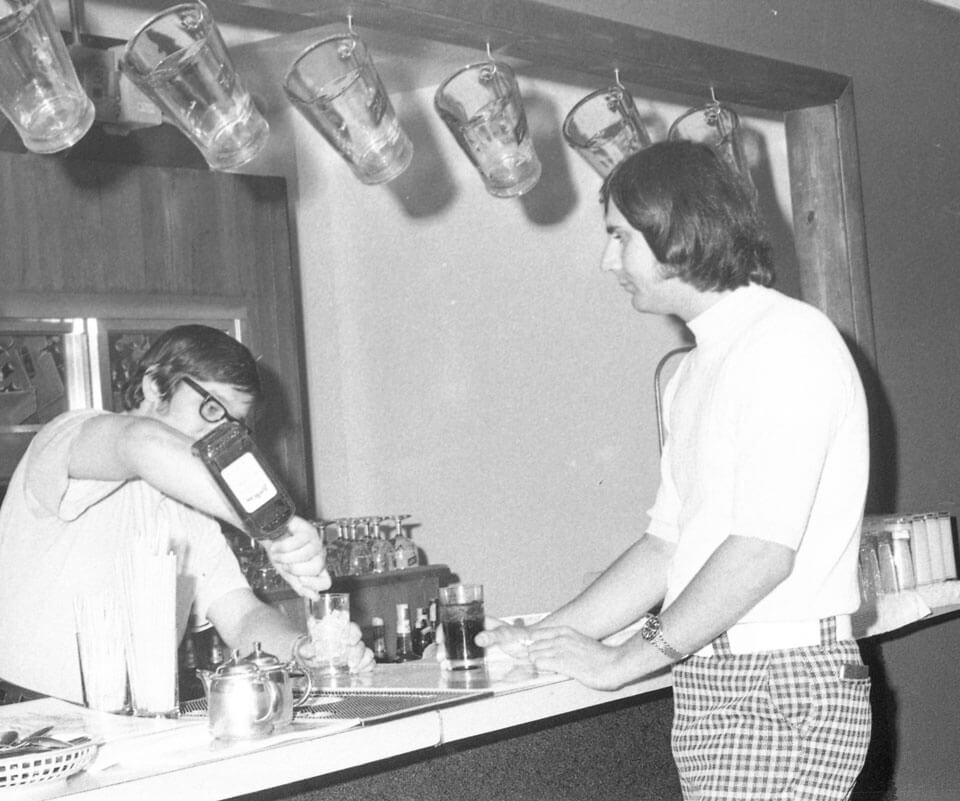 A bartender pouring a drink for a man at a bar, surrounded by hanging glasses and bar utensils.