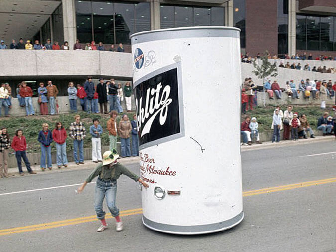 A large novelty Schlitz beer can costume moves down a street during a public event.