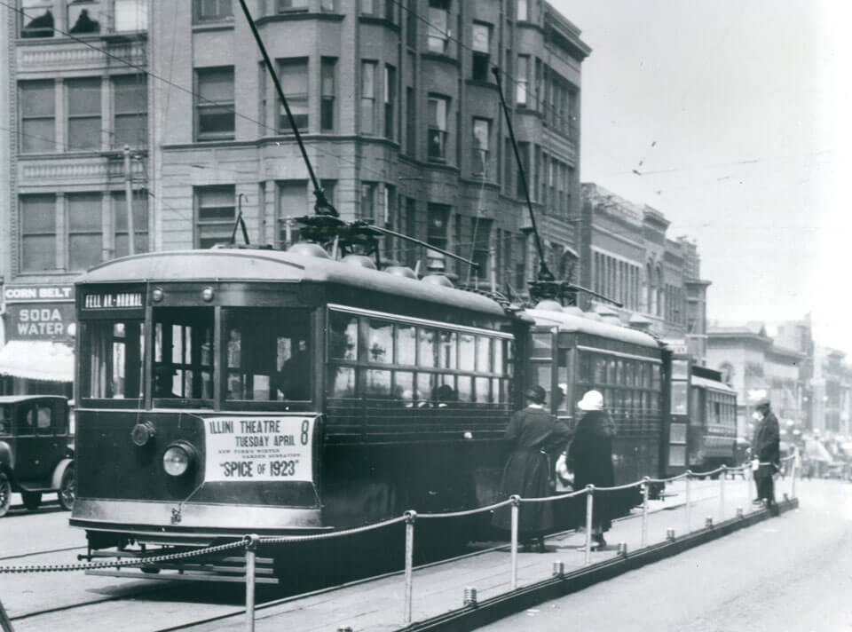 Historic streetcars on tracks with pedestrians nearby, set against a backdrop of early 20th-century brick buildings.