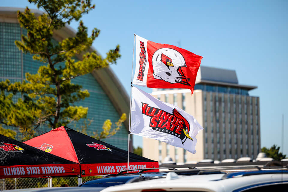 Flags of Illinois State University fly above tents, with buildings in the background.