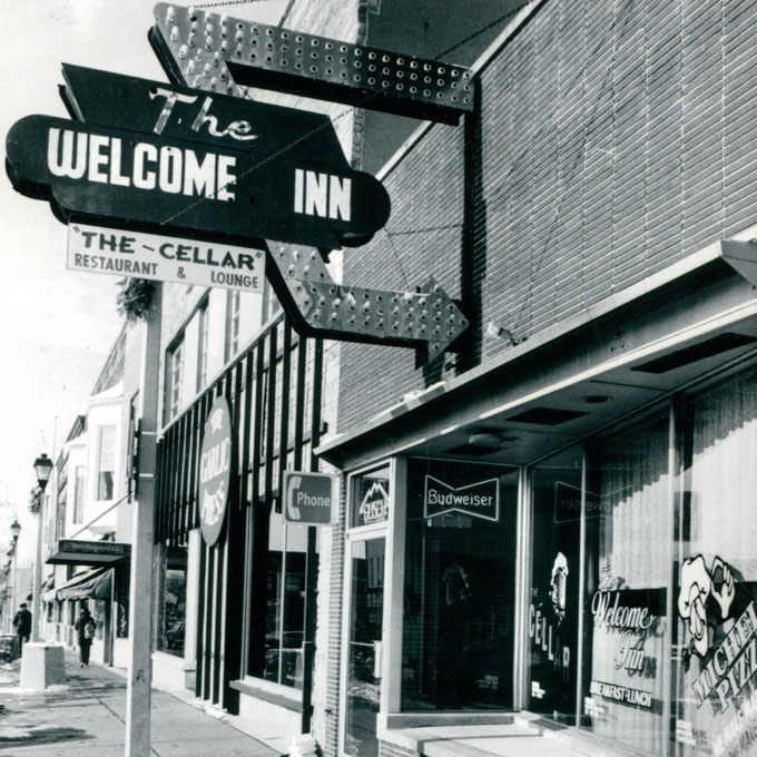 Street view of 'The Welcome Inn' with a vintage sign and storefront. Streetscape includes brick building and nearby businesses.