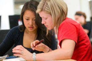 Two young women sit at a table and look intently at a document. It is clear they are working together.