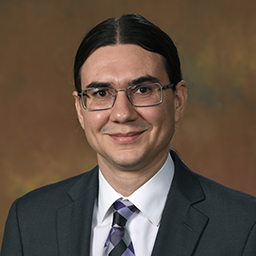 Rafael Copat, smiling, wearing glasses, a white collared dress shirt and tie, with a grey coat over
